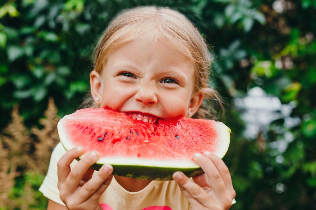 A little girl biting into a slice of watermelon.